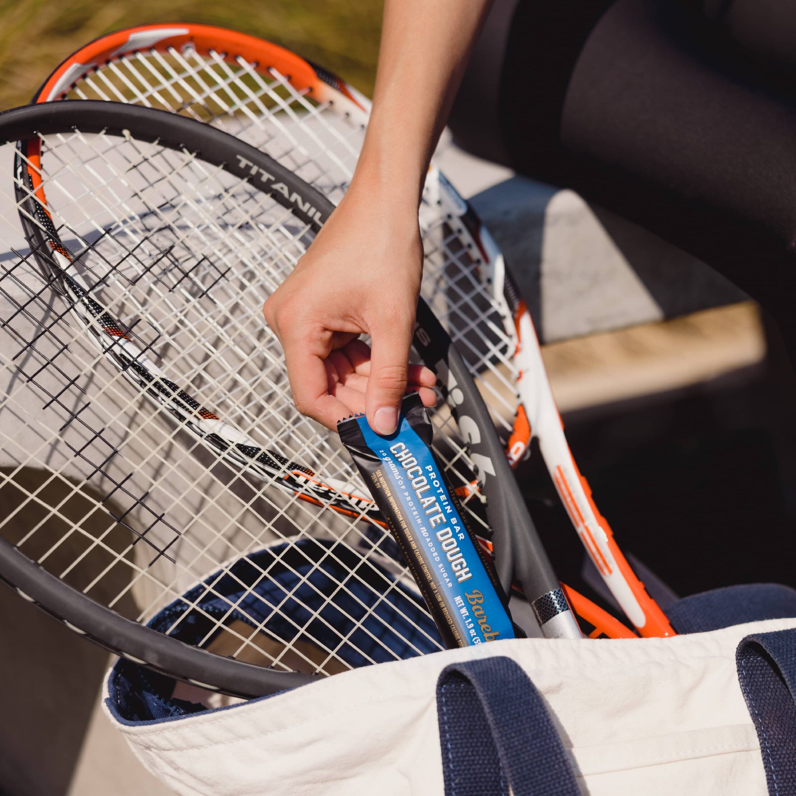 Hand retrieving a protein bar from a tennis bag with rackets.